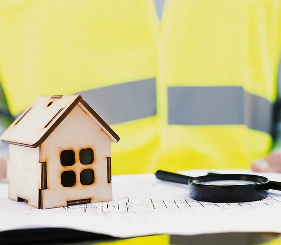 نموذج منزل خشبي صغير موضوع على مخطط هندسي مع عدسة مكبرة، يرتدي المهندس سترة سلامة صفراء في الخلفية.